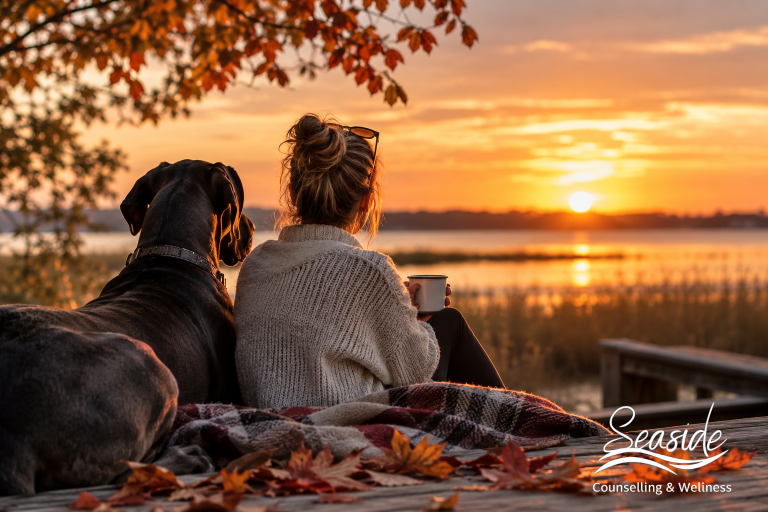 Woman sitting beside a black dog by the water during fall, representing seasonal mood changes and fall mental health